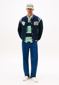 Giacca varsity navy con strisce bianche e ricami, polo a righe menta e navy sotto, jeans blu rilassati e sneakers bianche.