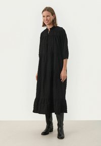 Schwarzes, langärmliges Kleid mit strukturiertem Muster, Rüschenausschnitt und gestuftem Saum. Kombiniert mit kniehohen schwarzen Stiefeln.