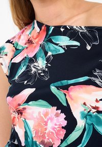 Zbliżenie ramienia kobiety w granatowej bluzce z dużymi różowymi, koralowymi i turkusowymi wzorami kwiatowymi oraz asymetrycznym dekoltem.