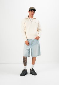 Man wearing beige knit sweater, light denim shorts, dark sneakers, and a checkered cap, standing with one hand in pocket against white background.