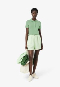 Polo-shirt vert, short rayé vert et blanc, portant une veste verte et un sac fourre-tout beige, avec des baskets blanches. Texture de tissu lisse.