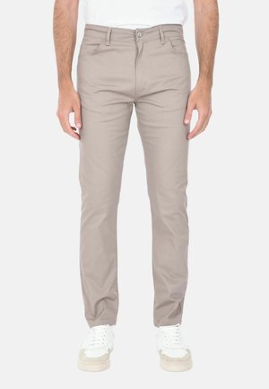 Uomo che indossa pantaloni beige aderenti e sneakers bianche, in piedi davanti a uno sfondo chiaro e uniforme.