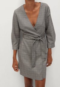 Szara w kratkę sukienka w stylu wrap z głębokim dekoltem w serek, trzy-czwartowymi rękawami i związanym w talii pasem. Materiał ma teksturowany, kratkowany wzór.