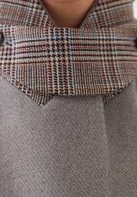 Šedý vlnený kabát s texturovaným povrchem, s hnědým a modrým károvaným límcem s vzorem. Výrazný úhlový střih na okrajích límce.