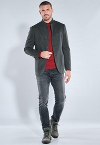 Gray jacket with black buttons, red sweater, gray jeans, and green boots. Model stands against a light backdrop, showcasing the outfit.