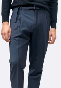 Pantaloni blu navy con una texture liscia, caratterizzati da passanti per cintura e una vita con fascia, pince anteriori e una vestibilità sartoriale, tenuti in vita da una cintura nera.