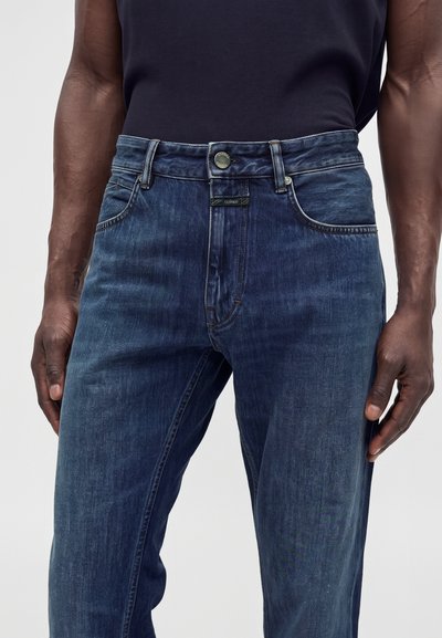 Homme portant un jean bleu foncé et une chemise noire, mains détendues sur les côtés, gros plan sur la taille et le haut des jambes.