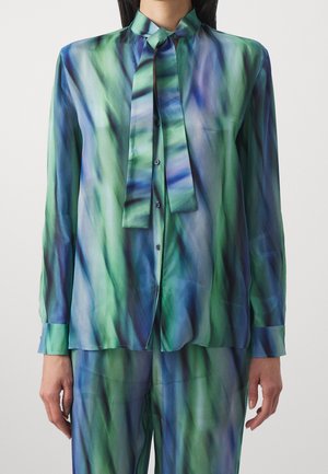 Woman wearing a long-sleeve button-up blouse and matching pants in green and blue abstract pattern with a tied neck scarf.