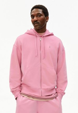 Mann trägt einen hellrosa Kapuzenpullover mit Reißverschluss und Kordeln sowie passende rosa Hosen, Hände in den Taschen, neutraler Hintergrund.