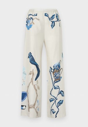 Femme portant un haut sans manches en satin bleu clair, un pantalon crème à jambes larges avec un imprimé floral et oiseau bleu, et des talons noirs ouverts à l'avant, debout avec une main dans la poche.
