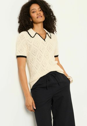 Frau mit lockigem Haar, die einen cremefarbenen Kurzarm-Pullover mit schwarzem Rand und schwarze Jogginghosen mit Kordelzug trägt, und vor einem weißen Hintergrund steht.