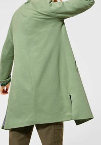 Cappotto verde a maniche lunghe con una forma leggermente svasata, realizzato in tessuto liscio, con fessure laterali e un piccolo dettaglio a anello al colletto.