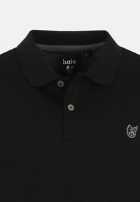 Mustapaita, jossa on kaulus, kaksinappinen etumus ja pieni brodeerattu logo vasemmassa rinnassa. Pehmeä kangas, jossa on teksturoitu kudonta.