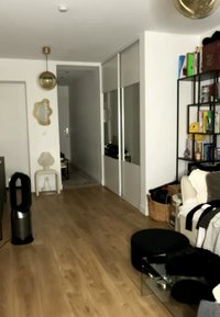 Soggiorno con pavimento in legno chiaro, pareti bianche, scaffali neri con libri, divano bianco, pouf rotondo nero e un corridoio con porte a specchio scorrevoli.