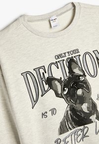 Grijze sweater met een zwart Franse bulldog graphic, met tekst in zwart en zilver. De stof heeft een gladde textuur en een ribbelhals.