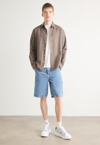 Světle hnědá košile na knoflíky, bílé tričko, modré denimové šortky a bílé tenisky s černými grafickými vzory. Stojící na neutrálním pozadí.