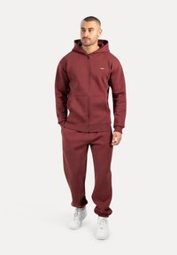 Bordová mikina s kapucí a tepláky vyrobené z měkkého fleecového materiálu, s jakostním zipem, předními kapsami a žebrovanými manžetami. Bílé tenisky dokreslují celý vzhled.