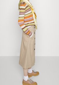 Pull à manches longues rayé en marron, orange et vert ; jupe beige à boutons ; écharpe en soie jaune ; chaussures à plateforme marron avec des chaussettes blanches.