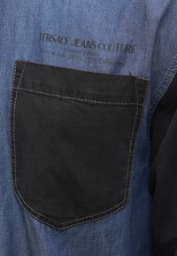 Koszula dżinsowa z niebieską górną częścią i czarną kieszenią, z widocznym przeszyciem. Napis brzmi: "Versace Jeans Couture, Edycja Limitowana."