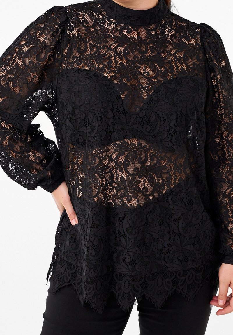 Blouse en dentelle noire à manches longues avec motifs floraux, épaules bouffantes et ourlet festonné portée sur un sous-vêtement noir.