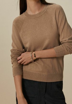 Femme portant un pull en maille couleur camel et un pantalon noir, montrant des bracelets en or et en chaîne sur son poignet gauche croisé devant son corps.