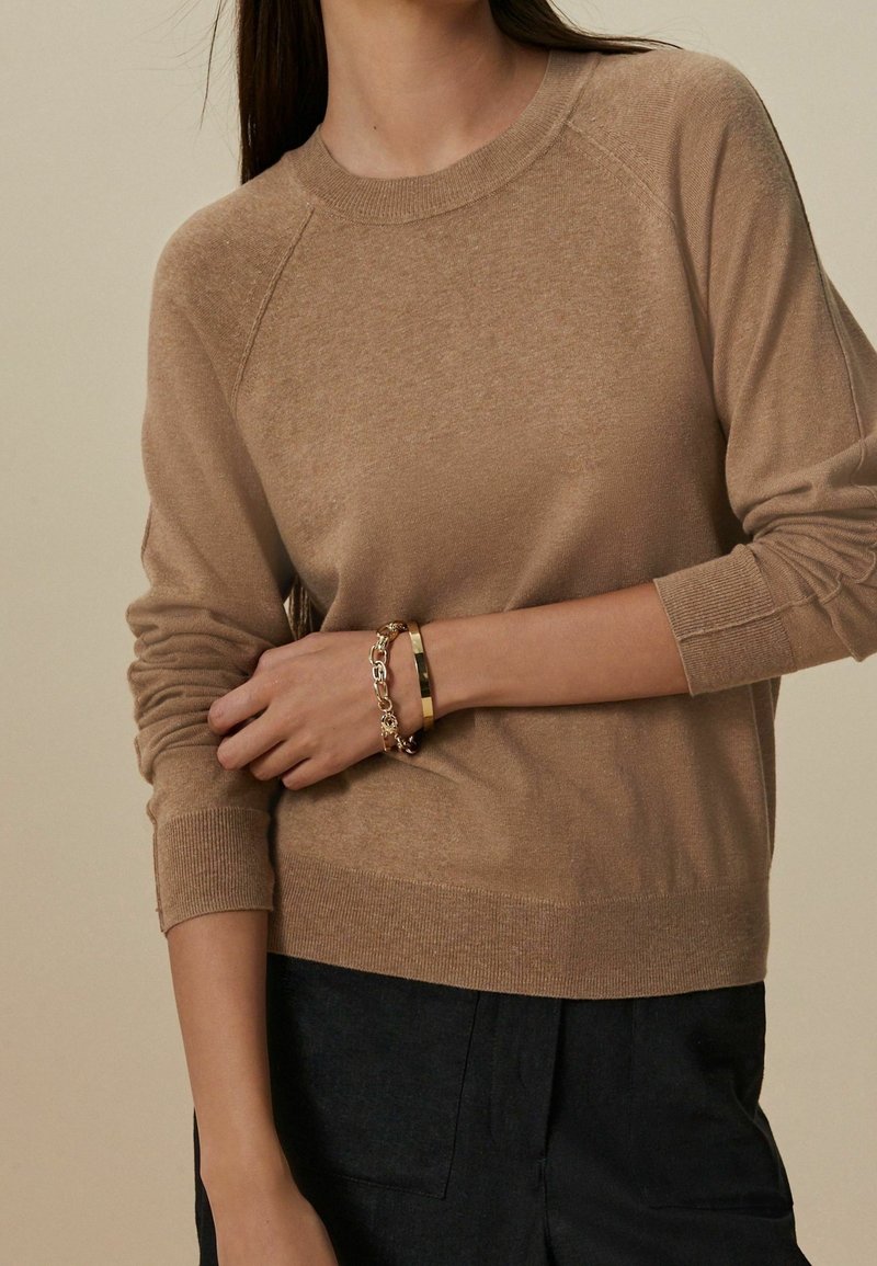Femme portant un pull en maille couleur camel et un pantalon noir, montrant des bracelets en or et en chaîne sur son poignet gauche croisé devant son corps.