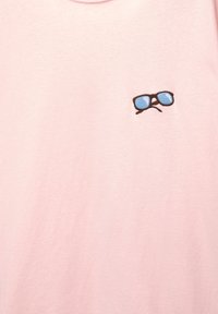 Tricou din bumbac roz deschis, cu un mic personaj brodat care poartă ochelari de soare albaștri și un contur maro aproape de zona pieptului.