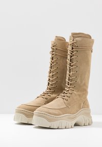 Botas de ante beige hasta la rodilla con cordones, suela gruesa texturizada y costuras reforzadas. Cuenta con ojales de metal y un acolchado en el cuello para mayor soporte.