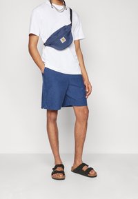 Bílá tričko, modré šortky a černé sandály. Modrý bum bag s logem je nošený přes hruď. Neformální a pohodlný outfit.