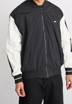 Homme portant une veste bomber noire et blanche zippée, avec des poignets et un ourlet côtelés, debout devant un fond clair et uni.