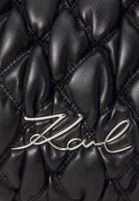 Melna ādas rokassoma ar quilted dizainu, kurā ir paceltas, noapaļotas zonas. Sudraba logo akcenta ar kursive rakstītu tekstu. 