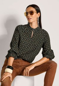 Schwarze Bluse mit goldenen, kreisförmigen Mustern, Schlüssel-Detail und gerafften Ärmeln, kombiniert mit braunen Hosen und auffälligen Armreifen.