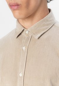 Beżowa koszula z korduroju z kołnierzykiem, listwą na guziki z białymi guzikami oraz subtelną, pionową fakturą prążków.