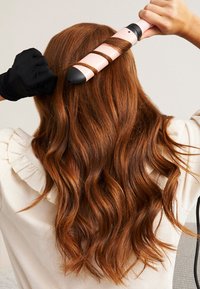 Haarglätter mit einer schwarzen Spitze und einem rosa Fass, der auf langen, gewellten rotbraunen Haaren verwendet wird. Der Benutzer trägt einen schwarzen hitzebeständigen Handschuh.