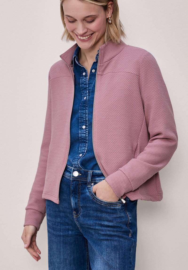 Frau trägt eine strukturierte, staubig-rosa Reißverschlussjacke über einem blauen Jeanshemd und Jeans, lächelt mit einer Hand in der Tasche.