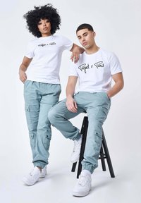 Twee personen dragen bijpassende witte t-shirts met zwarte logo's, in combinatie met lichtblauwe cargobroeken. De broeken hebben zakken en elastische boorden.