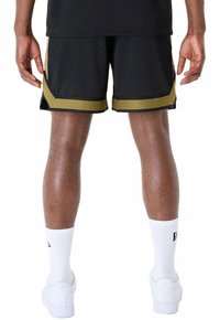 Pantalones cortos deportivos negros con acentos dorados, fabricados en tela de malla. Presentan un diseño con abertura lateral y se combinan con calcetines deportivos blancos y zapatillas.