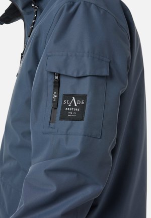 Navy blue waterproof jacket with a zip pocket and a patch label on the left shoulder. Smooth texture, modern design, and functional details.