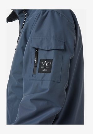 Navy blue waterproof jacket with a zip pocket and a patch label on the left shoulder. Smooth texture, modern design, and functional details.