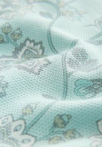 Teksturowana tkanina w jasnoniebieskim kolorze z kwiatowymi wzorami w odcieniach szarości i zieleni, charakteryzująca się miękkim, wyplatanym wyglądem oraz złożonymi detalami w projekcie.