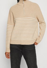 Beżowy sweter w prążki z półzamkiem, z poziomymi białymi paskami na klatce piersiowej i szerokimi rękawami. Miękka tekstura i luźny krój.