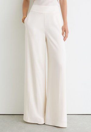 Crèmekleurige broek met wijde pijpen en hoge taille, gedragen door een persoon met één hand in de zak en een gouden armband om de pols.