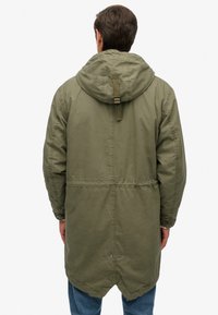 Olive green parka with a drawstring hood, elastic waist, and snap button closure at the cuffs, featuring a slightly curved hem.