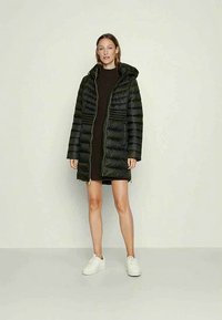 Schwarze Pufferjacke mit Kapuze, Reißverschluss und horizontalen Steppnähten. Lange Ärmel und mittellange bis zur Oberschenkel; kombiniert mit einem braunen Kleid und weißen Sneakers.