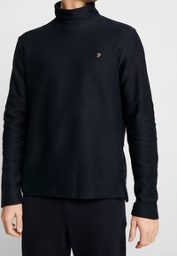 Granatowy pulower z teksturowanym wykończeniem, z wysokim kołnierzem, długimi rękawami, bocznymi rozcięciami i małym ikoną na piersi. Miękki materiał zapewnia luźny krój.