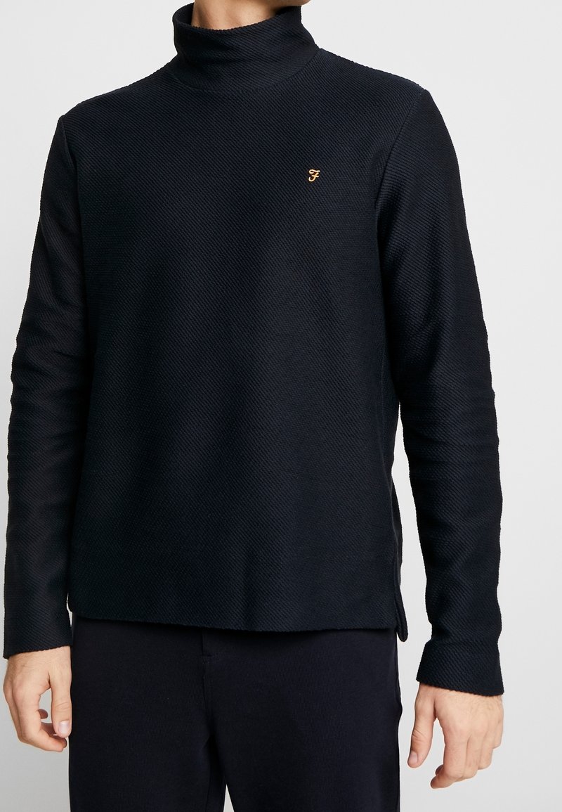 Granatowy pulower z teksturowanym wykończeniem, z wysokim kołnierzem, długimi rękawami, bocznymi rozcięciami i małym ikoną na piersi. Miękki materiał zapewnia luźny krój.