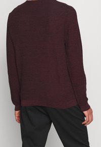 Vínový svetr s dlouhým rukávem z texturované látky, kulatým výstřihem a volným střihem, doplněný černými kalhotami, pohled zezadu.