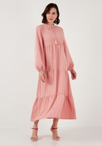 Robe maxi rose avec manches longues, niveaux froncés et fermeture à nouer. Le tissu a une texture douce. Assortie à des sandales multicolores.