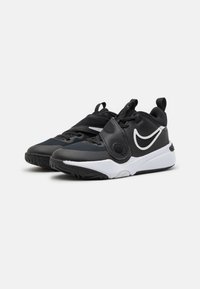Svarta och grå sportskor med en vit Nike-swoosh, utrustade med ett säkert spänne och texturerad sula, vinklade mot en enkel bakgrund.