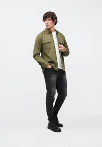 Olijfgroene blouse met knopen over een wit T-shirt, gecombineerd met donkergrijze spijkerbroeken en zwarte veterlaarzen. Model staat in een neutrale houding.